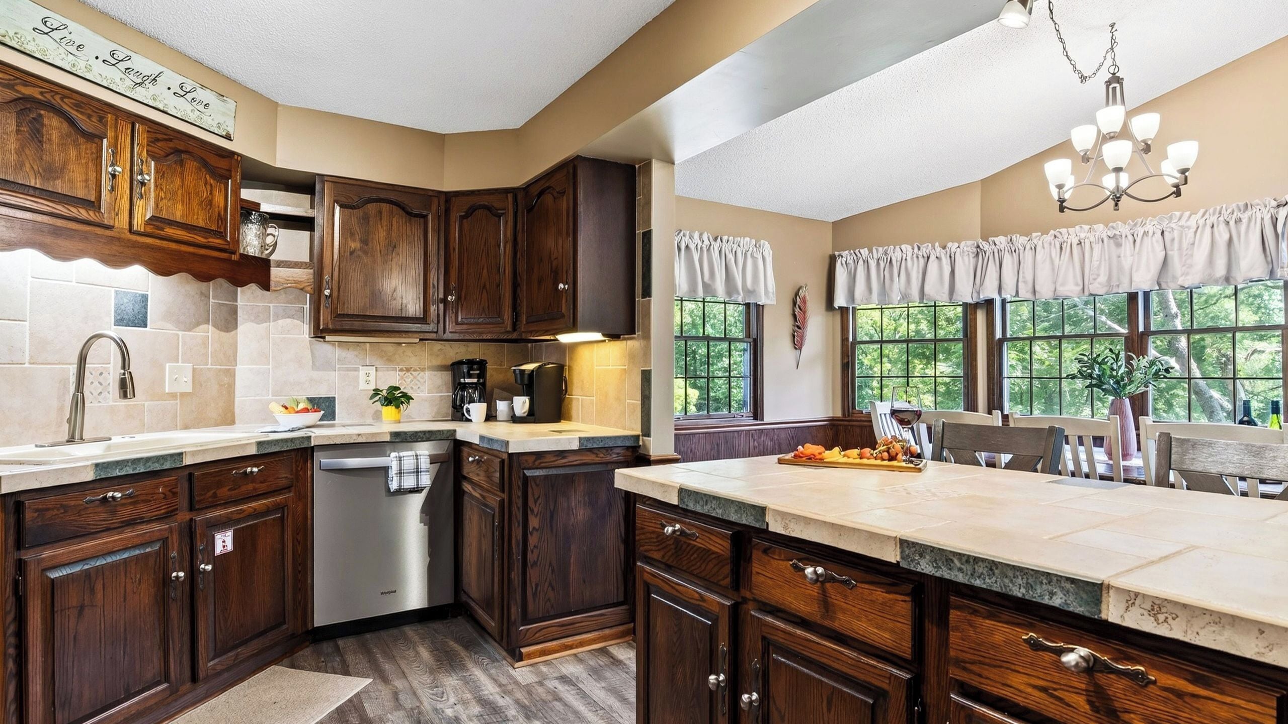 A kitchen with dark wood cabinets, a dishwasher, a coffee maker, and a dining area with a view of trees.