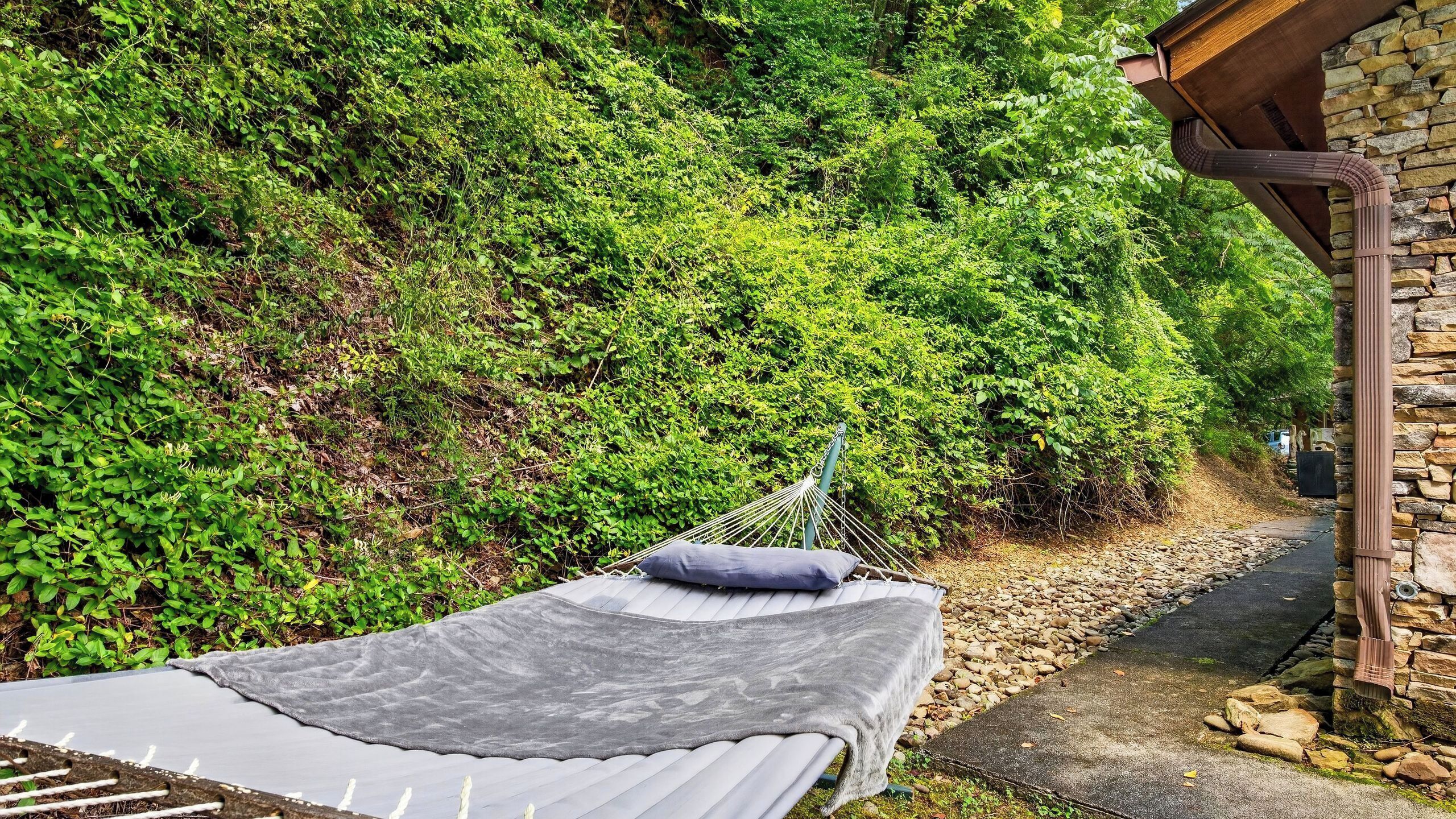 A hammock bed with a grey sheet and a blue pillow, set against a backdrop of dense green foliage and a stone wall.