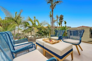 Eine Dachterrasse mit blauem Wicker-Möbel, einem Holztisch mit einer Obstschale und einer Flasche, und freier Aussicht auf Palmen und Gebäude.