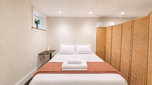 Ein Hotelzimmer mit Bett, Nachttisch, Wandklimaanlage und einem gewobenen Raumteiler.