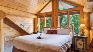 Ein Schlafzimmer einer Holzhütte mit einem großen Bett, zwei Nachttischen mit Lampen, einem Oberlicht und Blick auf den Wald.