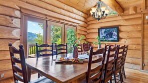 Comedor de cabaña de madera con una mesa dispuesta para seis personas, rodeada de sillas, una araña y un cuadro en la pared.
