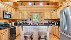 A modern kitchen with wooden cabinetry, stainless steel appliances, a central island with stools, and a window providing natural light. - Dreaming Bear Cabin by Avantstay Hot Tub Deck Game Room Soaking Tubs Near Nat'l Park Entry (Gatlinburg)