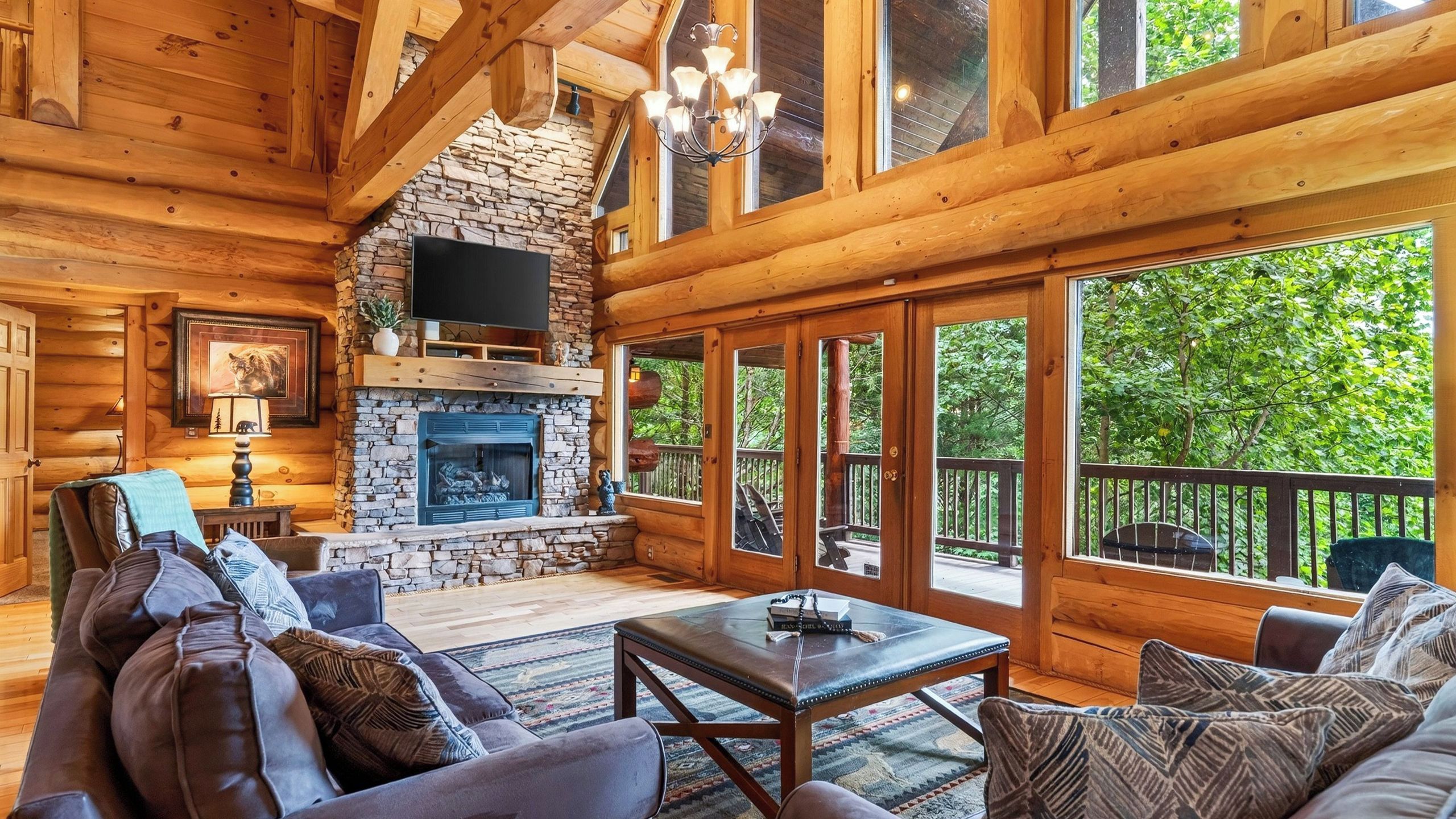 A log cabin interior with a stone fireplace, a television mounted above it, and a large window overlooking greenery.