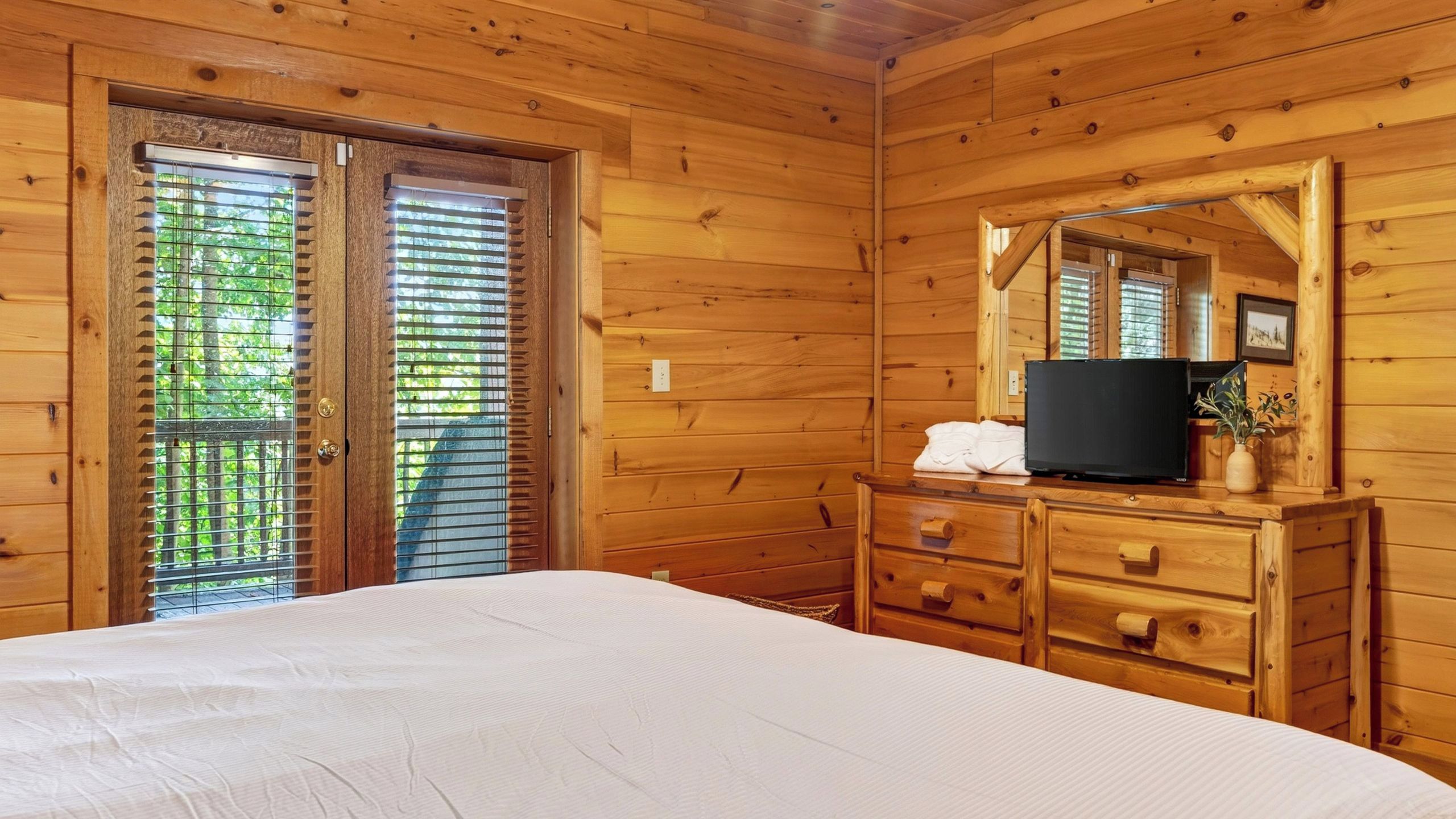 Habitación con paneles de madera, una cama, un aparador, un televisor y una ventana con persianas.