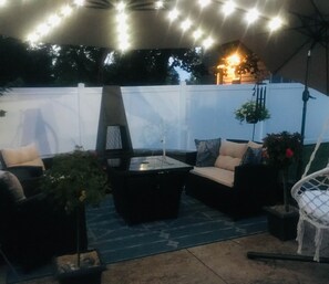 An outdoor patio with a black table, beige sofa, and a white chair. There are string lights overhead and a white fence in the background.