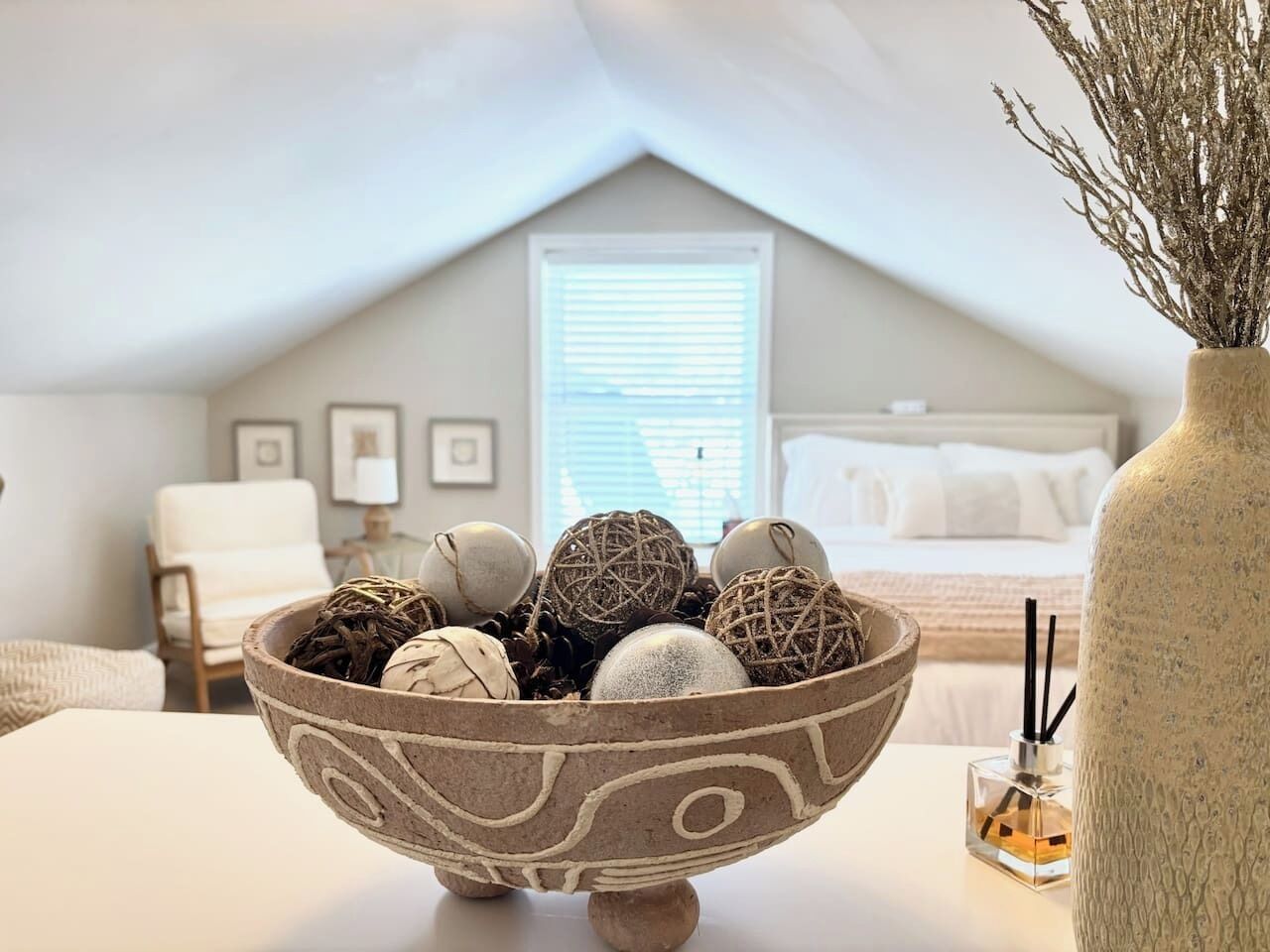 A bedroom with a sloped ceiling, a bed with white bedding, a bedside table with a decorative bowl, and a window with blinds.
