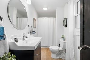 Un baño con lavamanos blanco, espejo e inodoro. Hay un perchero con una toalla y una planta pequeña sobre el lavamanos.