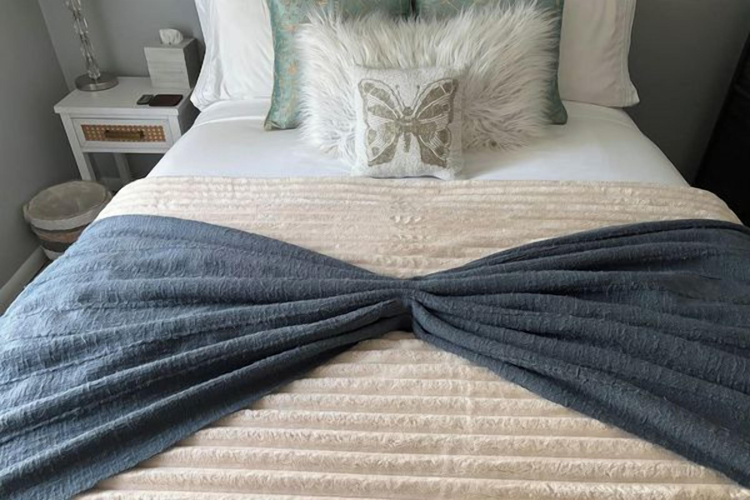 Una cama con una colcha azul, almohadas blancas y un cojín decorativo con diseño de mariposa.