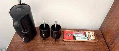A wooden table with a black coffee maker, two black mugs, and a tray with tea and milk cartons.