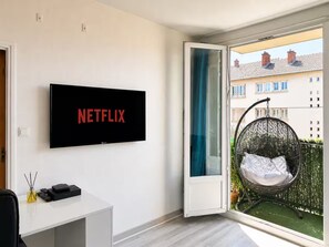 Ein modernes Wohnzimmer mit einem wandmontierten Flachbildfernseher, einem Schreibtisch mit Computer und Lampe sowie einem großen Fenster mit Blick auf einen Balkon und angrenzende Gebäude.