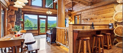 A wooden cabin interior with a dining area, bar stools, and a view of mountains.