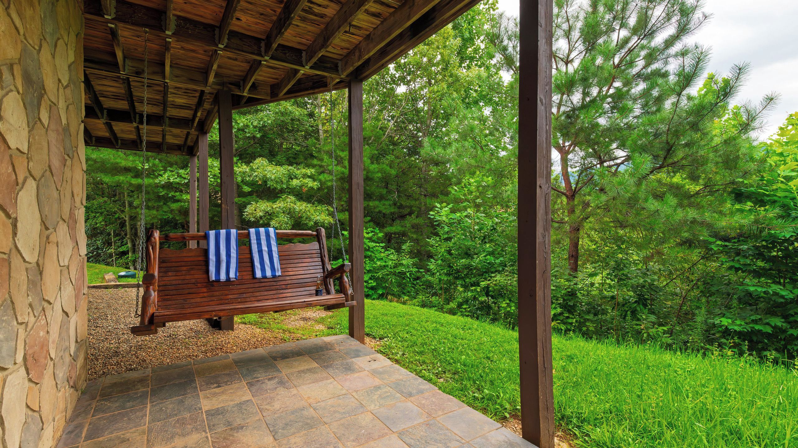 A wooden swing bench with towels, set on a stone-tiled patio with a view of a lush green forest.
