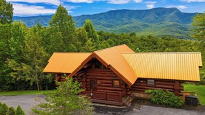 Eine Blockhütte mit geneigtem Dach, umgeben von üppigem Grün und Bergen im Hintergrund.