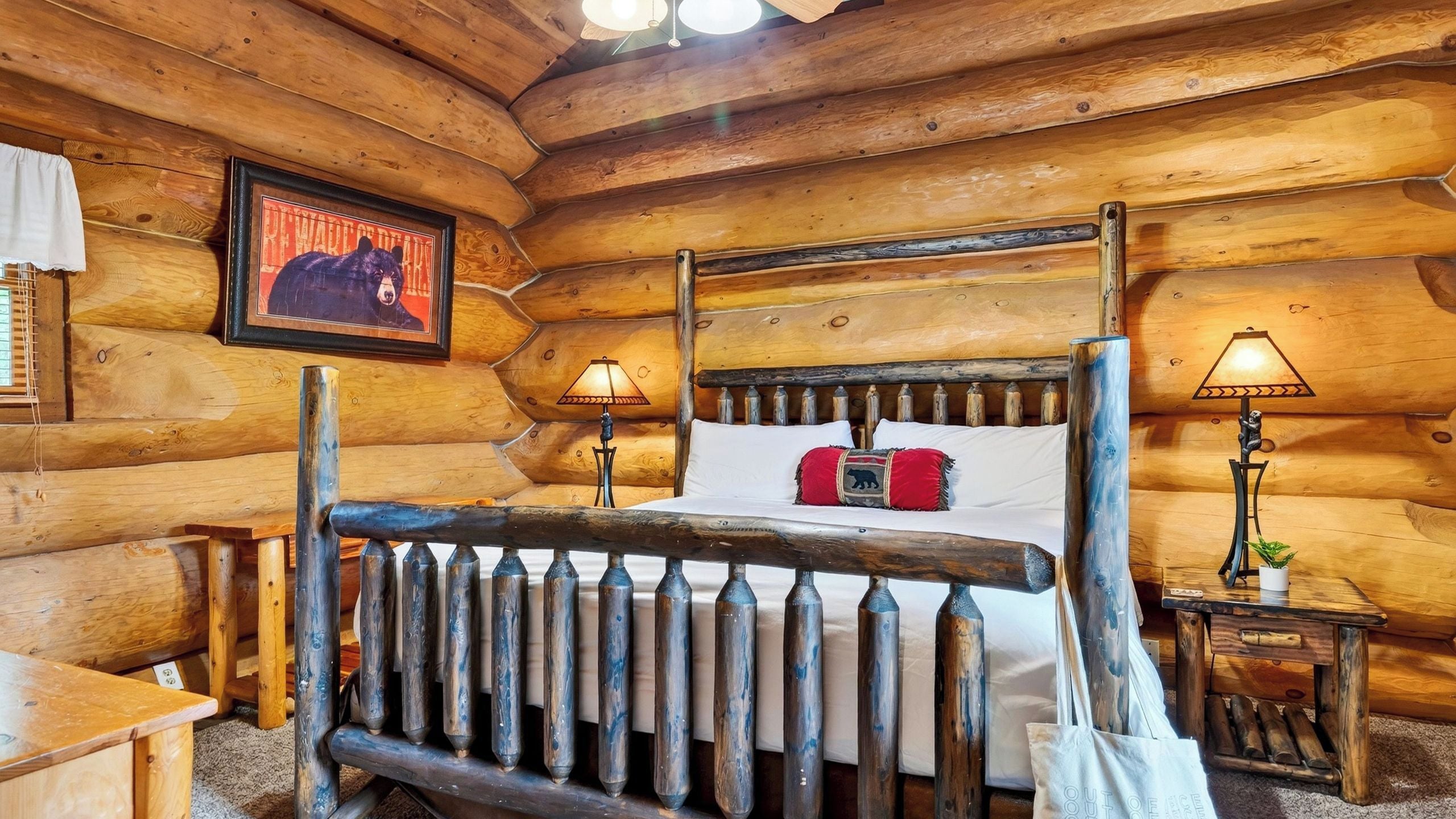 A log cabin bedroom with a four-poster bed, two bedside lamps, a framed bear picture, and a small table with a plant.