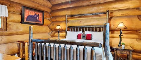 A log cabin bedroom with a four-poster bed, two bedside lamps, a framed bear picture, and a small table with a plant.