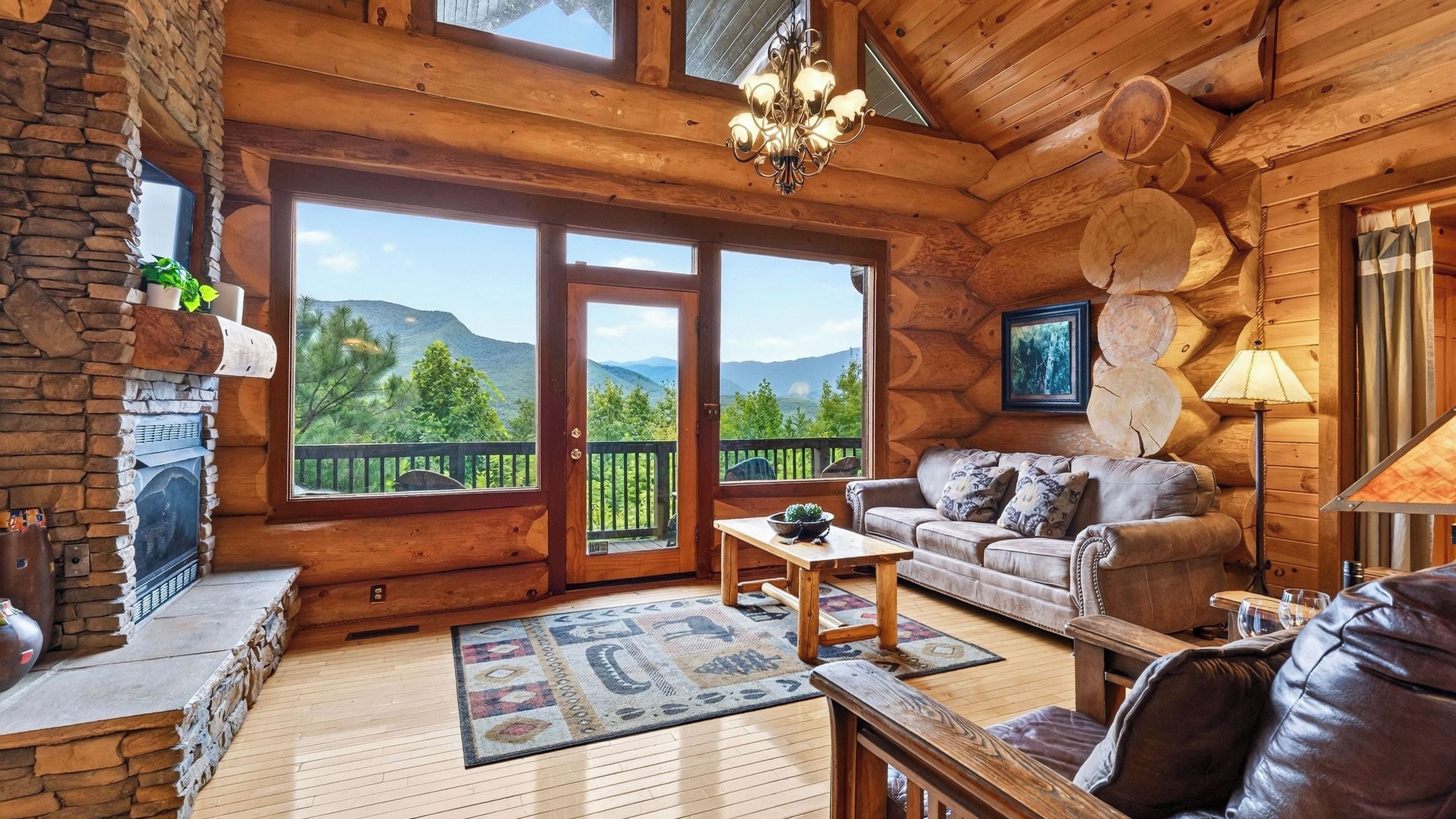 L’intérieur d’une cabane en rondins, avec une cheminée en pierre, un canapé, une table basse et une vue sur les montagnes par de grandes portes vitrées.