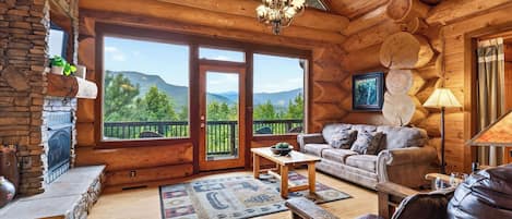 A log cabin interior with a stone fireplace, a sofa, a coffee table, and a view of mountains through large glass doors.