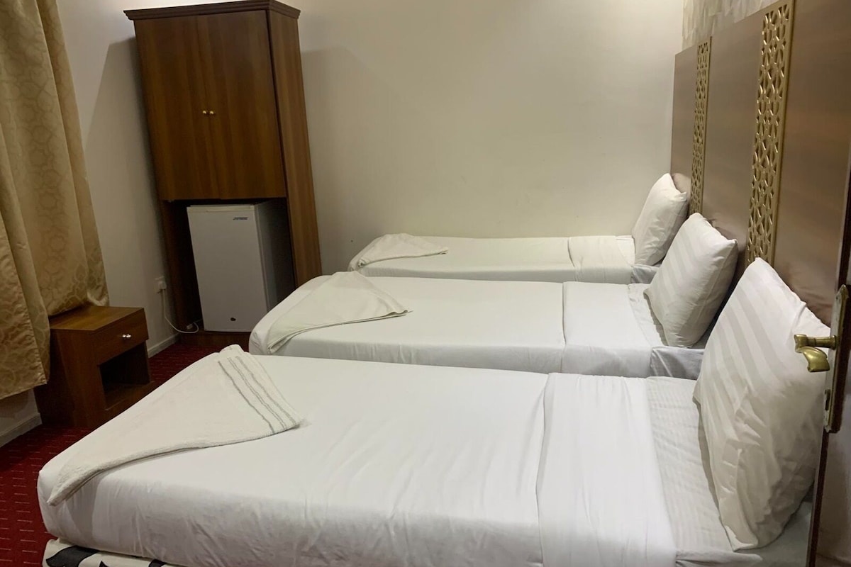 A hotel room with two single beds, a wooden wardrobe, a small refrigerator, and a red carpet.