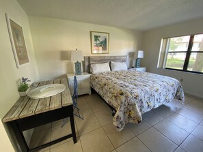 Een slaapkamer met een bed, een houten tafel, een nachtkastje, een lamp en een raam.