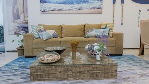 A living room with a sofa, decorative pillows, a glass coffee table with various items, and a painting of a sailboat on the wall. - Pousada Casa da Adriana (Fortaleza)