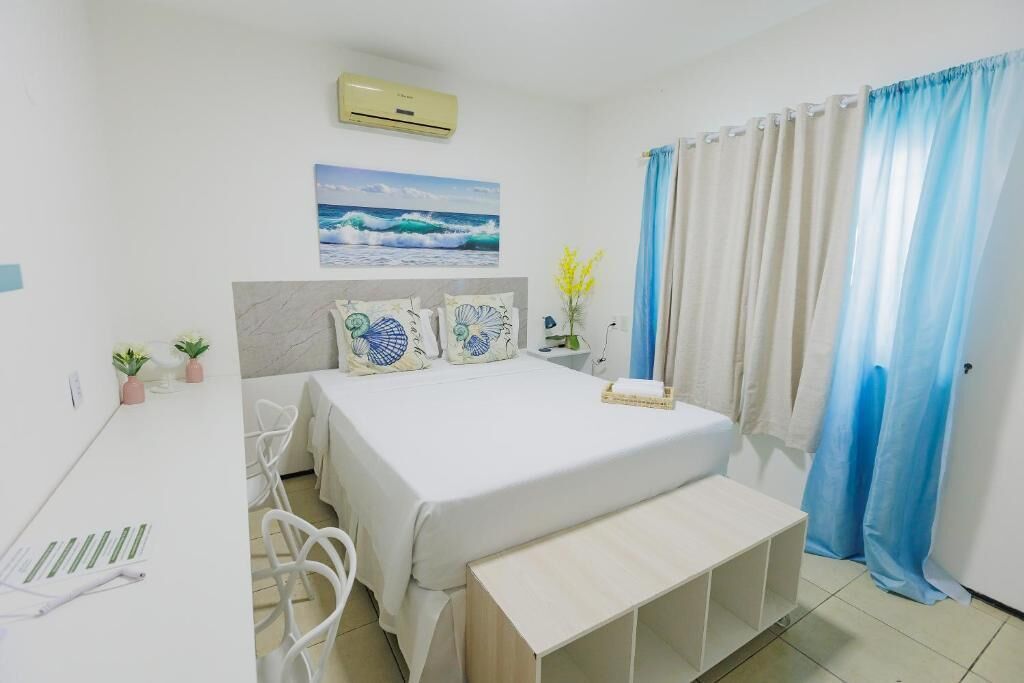 Un dormitorio con cama, escritorio, silla, mesita de noche, aire acondicionado mural y ventana con cortinas.