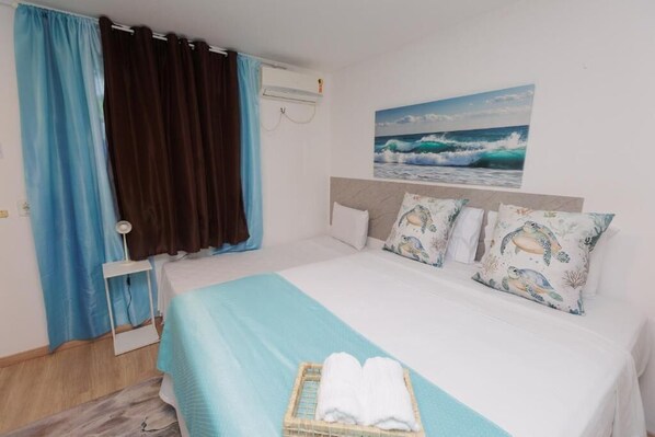 A hotel room with a bed, curtains, a wall-mounted air conditioner, and a painting of a wave.