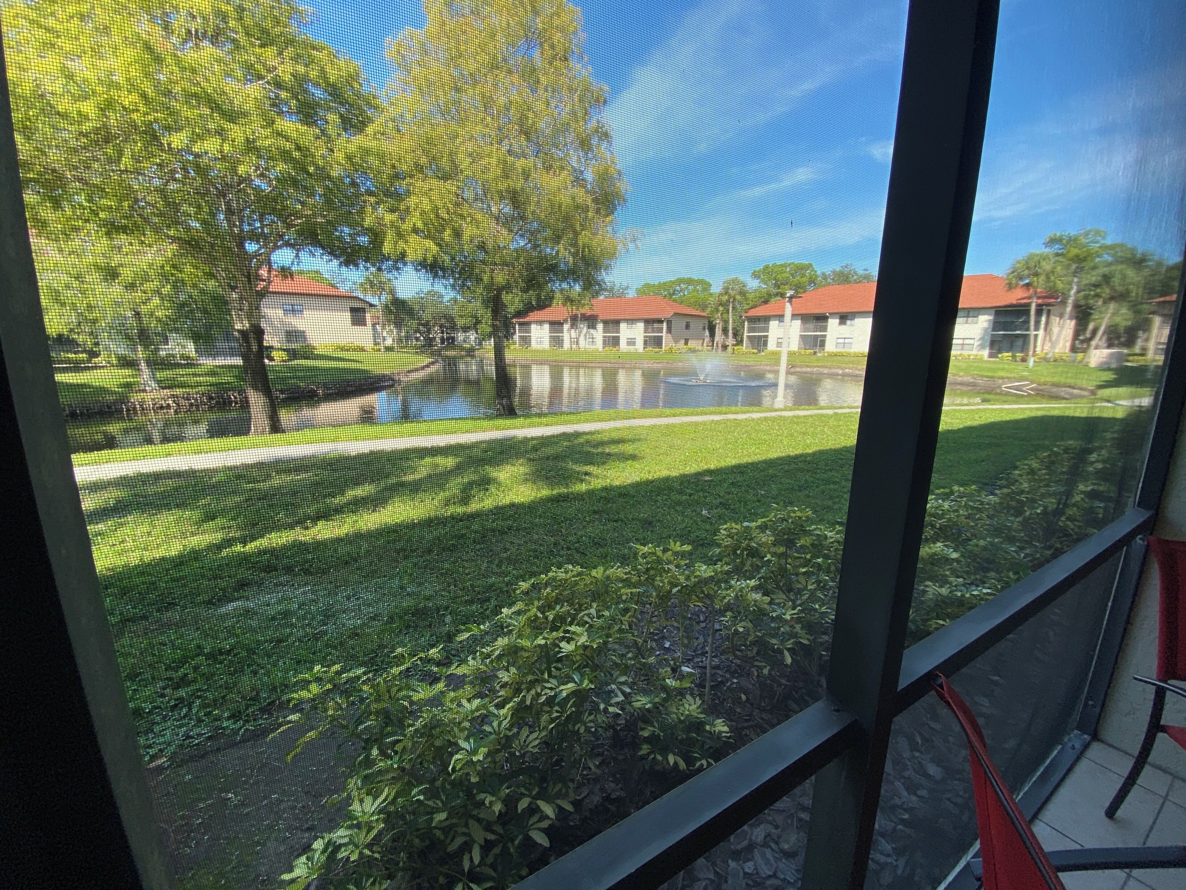 A view through a window with a mesh screen, displaying a grassy area with trees and a building with a red roof in the background.