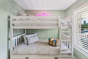 Una cama litera con un letrero de neón con forma de ola encima, una ventana con persianas, y una cesta de objetos en la cama inferior.