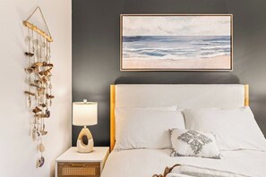 A bedroom with a bed, a bedside table with a lamp, a wall hanging, and a framed picture of the ocean. - Shore Cottage | Walk to Beach & Town | Patio Oasis (Belmar)