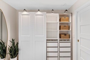 Un armario vestidor con puertas, estantes y cestas blancos, iluminado por luces de techo.