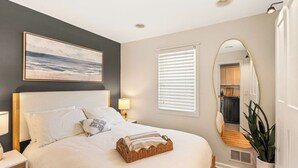 A bedroom with a large bed, a nightstand, a lamp, a wall-mounted painting, a window with blinds, and a mirror.