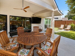 An outdoor patio with a round wooden table and chairs, a ceiling fan, and a TV mounted on the wall. - Settle in and stay awhile at this beautifully furnished luxury home! (New Braunfels)