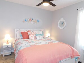 Ein Schlafzimmer mit einem Bett, zwei Nachttischen, einem Deckenventilator, Wandkunst und einem Fenster mit Vorhängen.