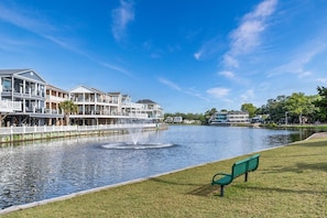 A waterfront property with a water fountain, a grassy area with a bench, and multi-story buildings with balconies. - Ocean Lakes 1576 Spacious 4BR with Hot Tub & Golf Cart (Myrtle Beach)