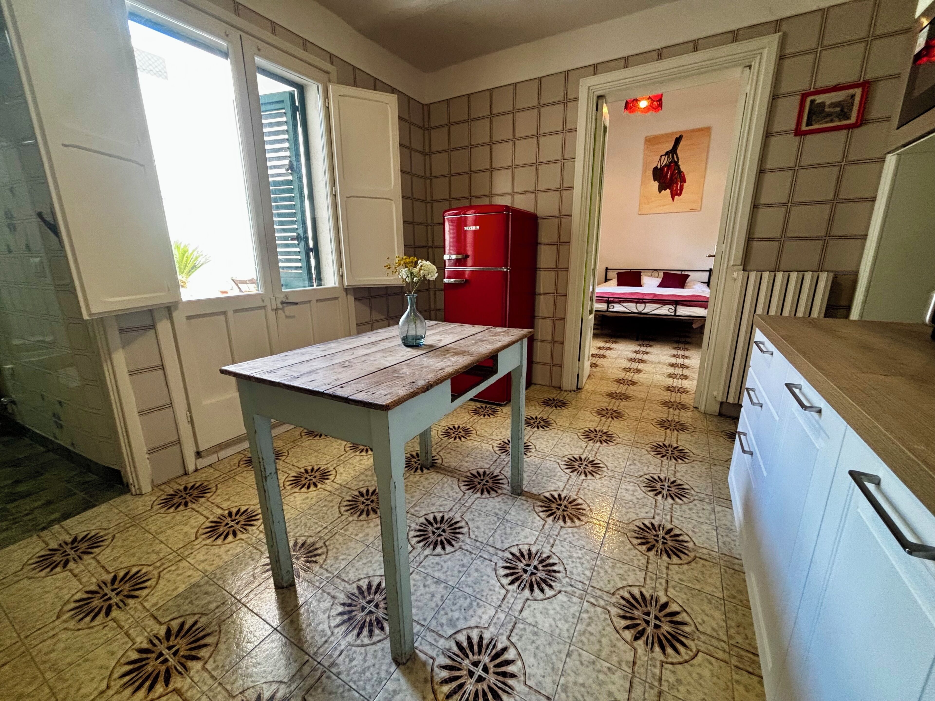 Una cucina compatta con pavimento piastrellato, un tavolo di legno, un frigorifero rosso e un letto sullo sfondo.