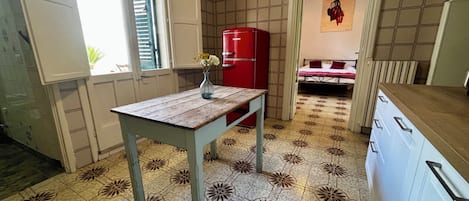 Cocina compacta con piso de baldosas, una mesa de madera, un refrigerador rojo y una cama al fondo.