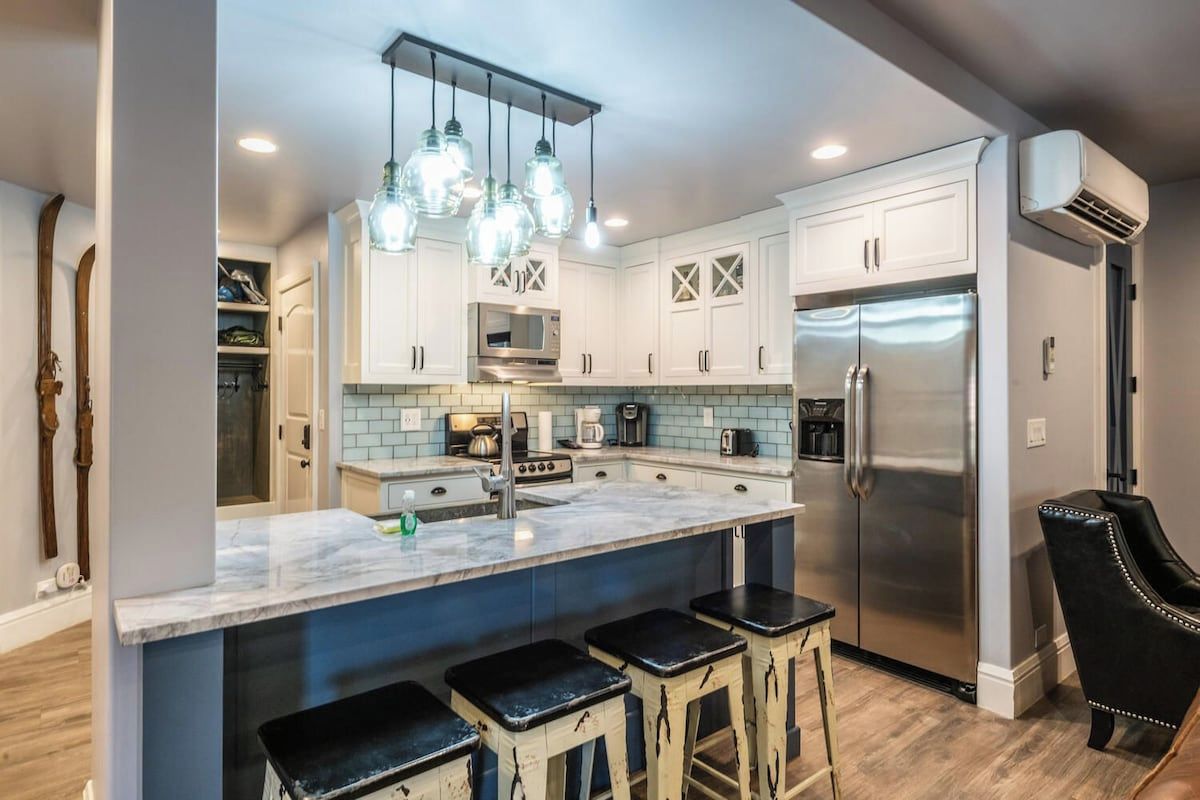 A modern kitchen with white cabinets, a stainless steel refrigerator, and a marble island with bar stools.