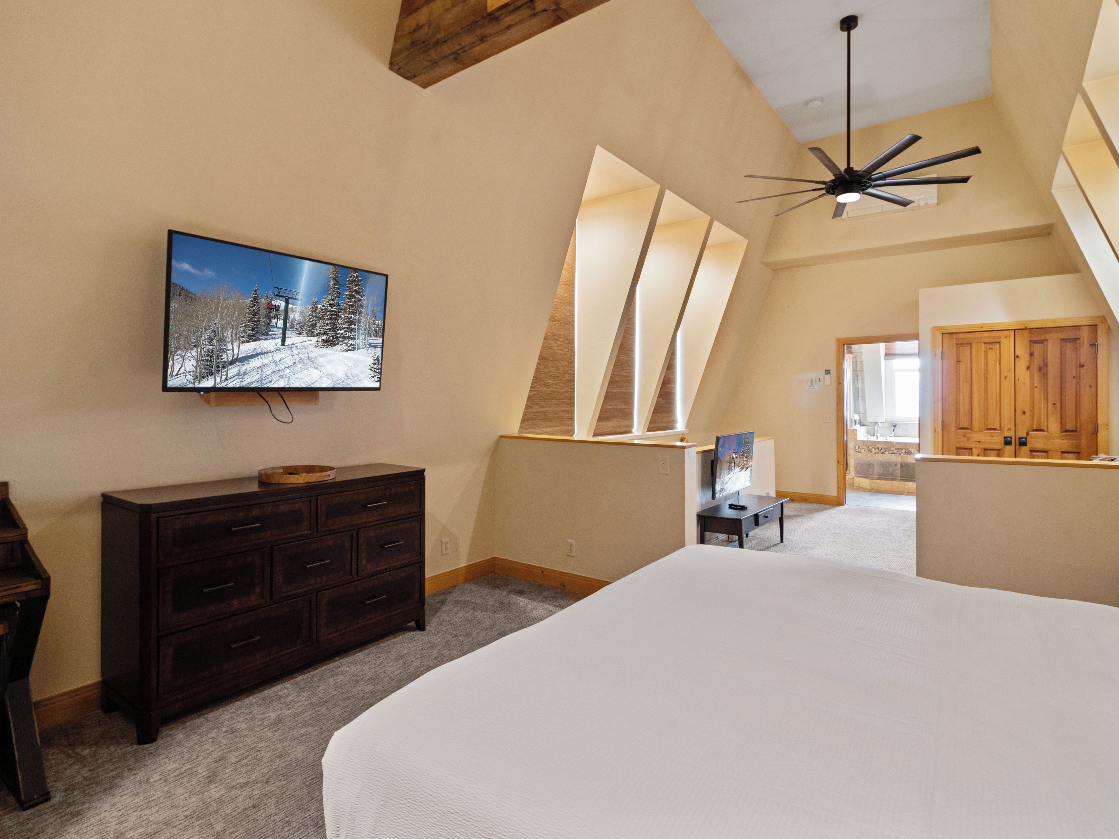 A bedroom with a large bed, a dresser, a TV mounted on the wall, and a ceiling fan.