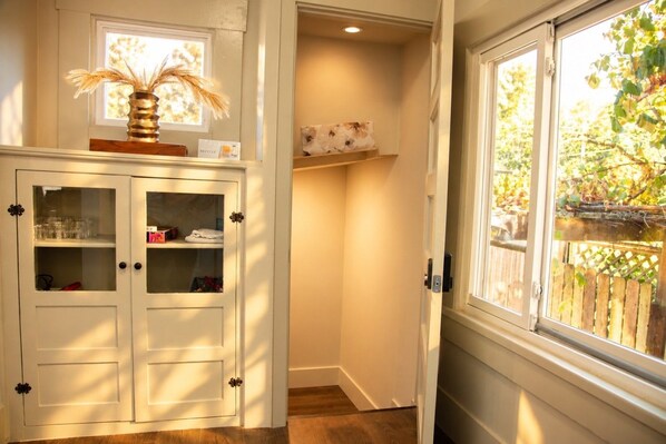 A room with a white cabinet with glass doors, a window with a view of greenery, and a shelf with decorative items.
