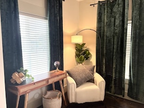 A corner room with a white armchair, a wooden side table with a globe and a plant, a lamp, and a window with blinds.