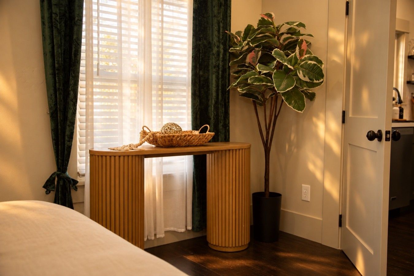 A room with a bed, a table with a basket, a potted plant, and a door leading to a kitchen.