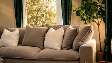 Ein beiges Sofa mit vielen Kissen, platziert in einem Raum mit einem großen Fenster und einem Blumentopf.