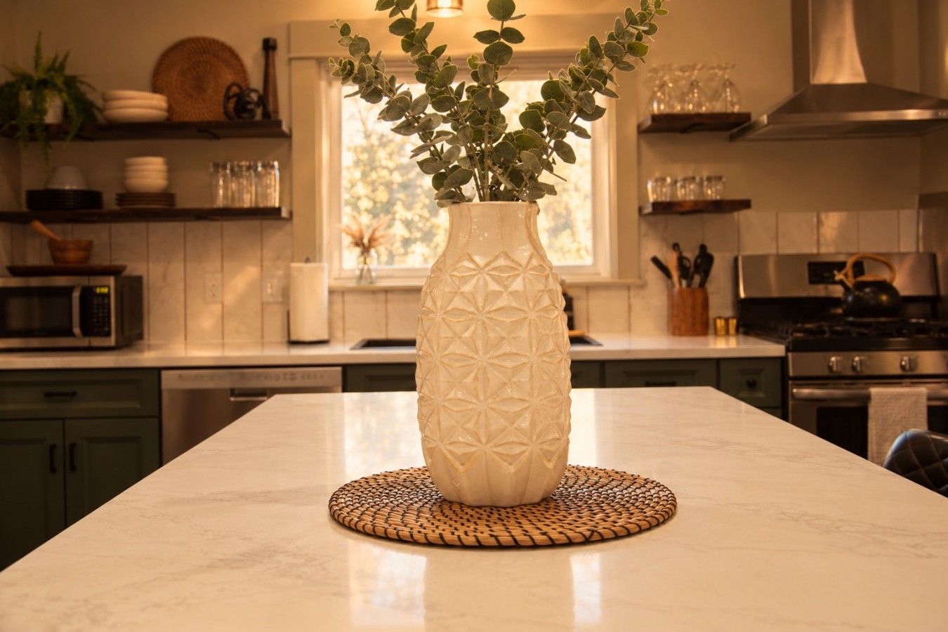 Une cuisine avec un vase blanc posé sur un plan de travail en marbre, entouré d’appareils de cuisine et d’une fenêtre.