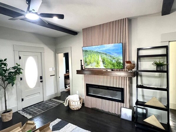 A modern living room with a flat-screen TV mounted on a wooden wall, a fireplace, a ceiling fan, and a black shelving unit. - The Moody Modern*2KING beds*2bath*Walkable* (Albany)