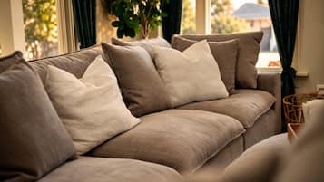 A beige sofa with multiple cushions, positioned near a window with curtains, and a plant in the background.