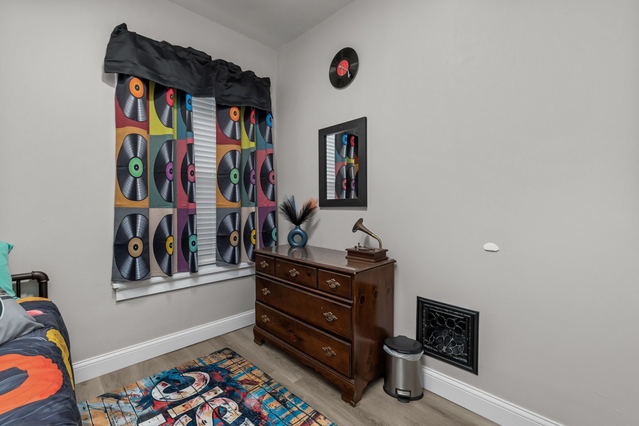 Un dormitorio con una ventana cubierta por una cortina con estampado de disco de vinilo, un aparador, un radiador y un reloj en la pared.