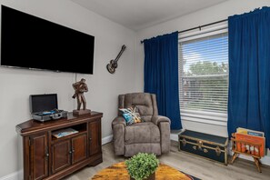 Ein Wohnzimmer mit Flachbildfernseher, Gitarre, Liegesessel, Holzschrank, Pflanze und einem Fenster mit Vorhängen.