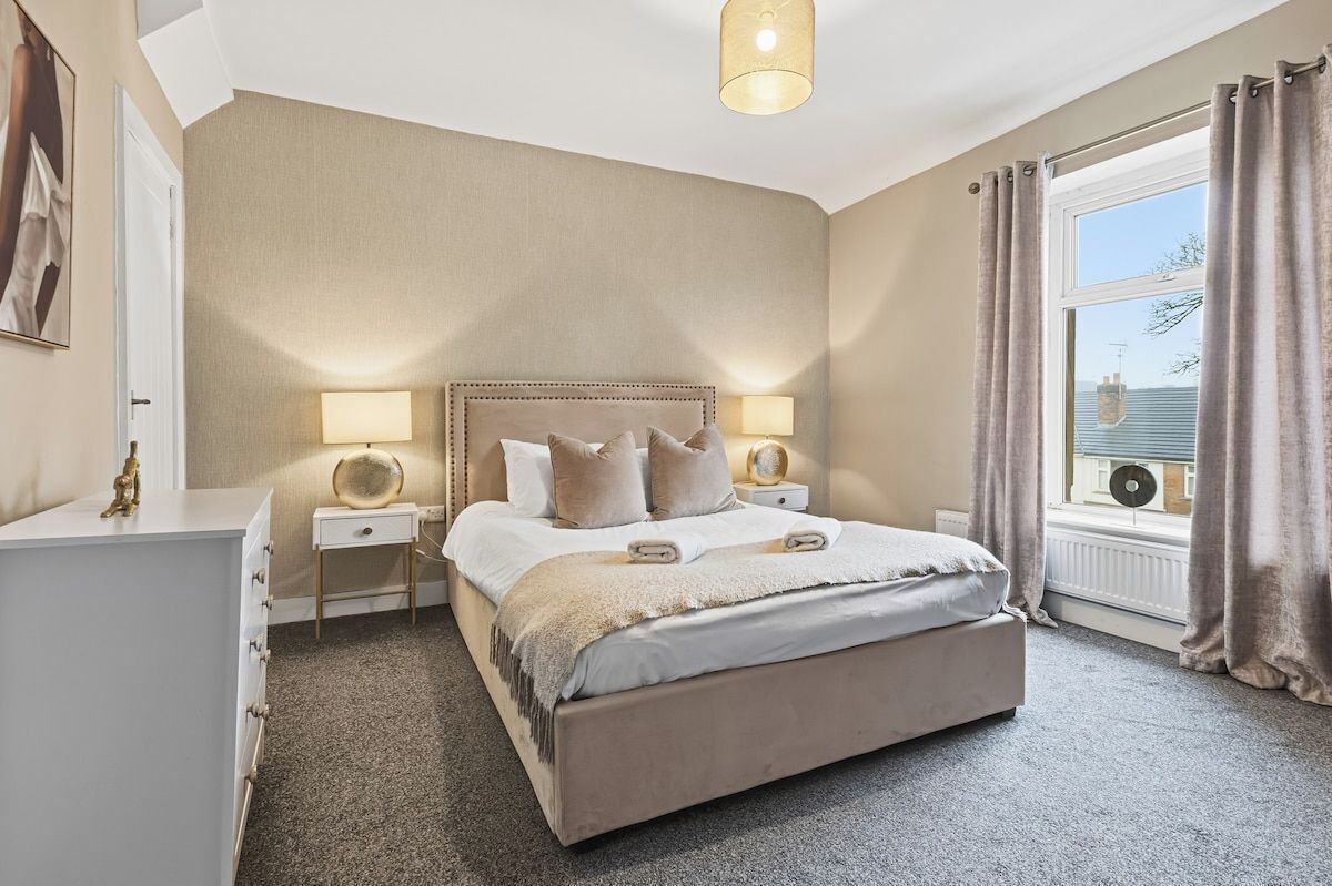 Ein Schlafzimmer mit einem großen Bett, Nachttischlampen, einem Nachttisch, einer Kommode und einem Fenster mit Vorhängen.