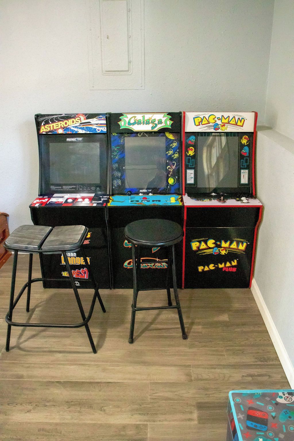 Three arcade machines, two stools, and a small table in a room with wooden flooring.